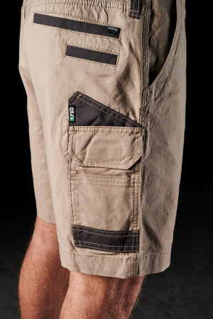 Close-up of a person's legs wearing the FXD WS-3 Shorts in beige, featuring black trim details and multiple utility pockets. Crafted from stretch cotton canvas for durability, these shorts sport a rugged look with reinforced triple needle seams and include a small pocket labeled "CAT." The background is black and indistinct.