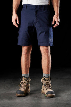 A person is standing against a dark background, wearing the rugged FXD WS-3 Short in navy blue, paired with gray socks and tan hiking boots. The bottom half of their body is visible, showcasing the resilience and durability of the stretch cotton canvas shorts.