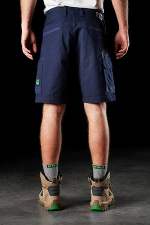 A person stands with their back to the camera, wearing the FXD WS-3 Short in dark blue, made of stretch cotton canvas for comfort and durability, grey FXD socks, and beige hiking boots with green soles. The background is black, and the floor appears to be concrete.