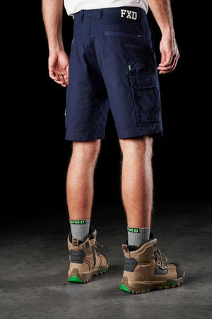A person stands facing away, wearing FXD WS-3 shorts in navy blue, made from stretch cotton canvas with the "FXD" logo visible on the back. They have on light gray socks and sturdy brown work boots featuring green accents. The dark background highlights the lower half of their body, showcasing both comfort and durability.
