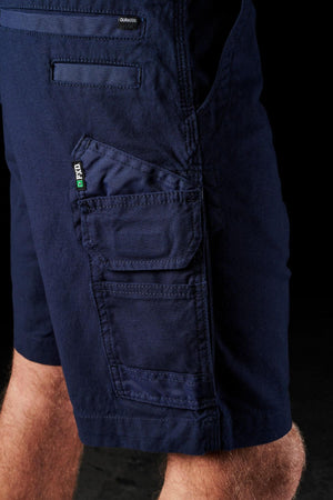 Close-up of a person wearing FXD WS-3 Shorts in dark blue. Crafted from stretch cotton canvas, these shorts boast multiple pockets with flaps and a small label on one pocket. The person's lower thigh and knee are visible, highlighting the comfort and durability embedded in every stitch. The background is black.