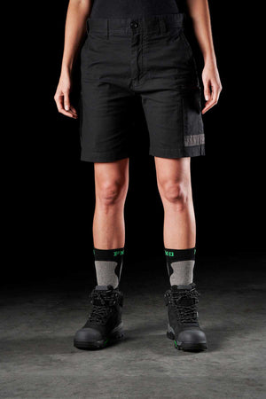 A person wearing the FXD Womens WS-3W Shorts in black, featuring DURA500 reinforced pockets, stands against a dark background. Black and grey socks with green accents peek out from sturdy black work boots. The shot is from the waist down, perfectly showcasing the durable and stylish stretch work shorts and footwear.