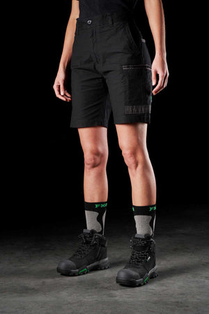 A person stands against a dark background, wearing FXD Women's WS-3W Shorts with DURA500 reinforced pockets, black and green hiking boots, and black and green patterned socks. Only their legs and part of their torso are visible.