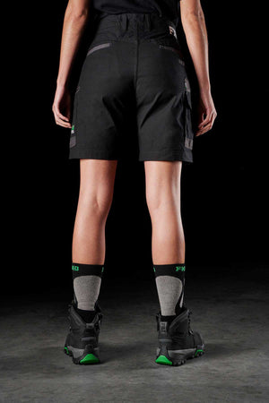 A person stands facing away from the camera, wearing FXD Womens WS-3W Shorts with DURA500 reinforced pockets, gray and black socks, and black boots with green accents. The background is dark, and the legs are partially visible from the calves down to the feet.