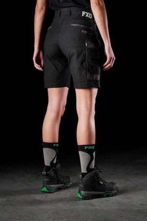 A person stands with their back facing the camera, wearing FXD Women's WS-3W Black Stretch Work Shorts featuring DURA500 reinforced pockets, a black shirt, black and green socks with "FXD" written on them, and black work boots with green accents. The background is dark and plain.