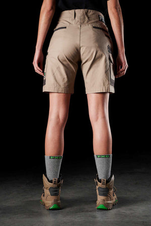 A person stands facing away, wearing FXD's Womens WS-3W khaki utility shorts with DURA500 reinforced pockets, a black top, gray socks with green patterns, and brown hiking boots with bright green soles. The background is a dark, neutral setting.