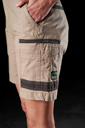 A person wearing tan FXD Womens WS-3W Shorts with their hand in the right pocket. The shorts feature black accents on the seams and edges, plus DURA500 reinforced pockets. The FXD logo is visible on the side pocket in this black background setting.