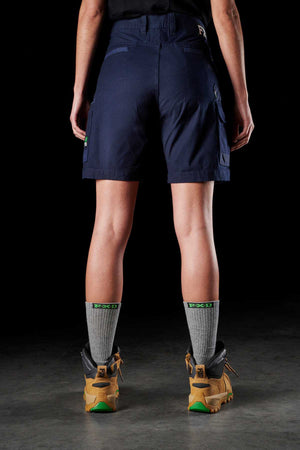 A person stands with their back to the camera, wearing the FXD Womens WS-3W Short in dark blue stretch fabric, featuring DURA500 reinforced pockets. They are also sporting a black shirt, grey socks, and tan hiking boots. The dark background enhances the contrast of their sturdy attire and footwear.