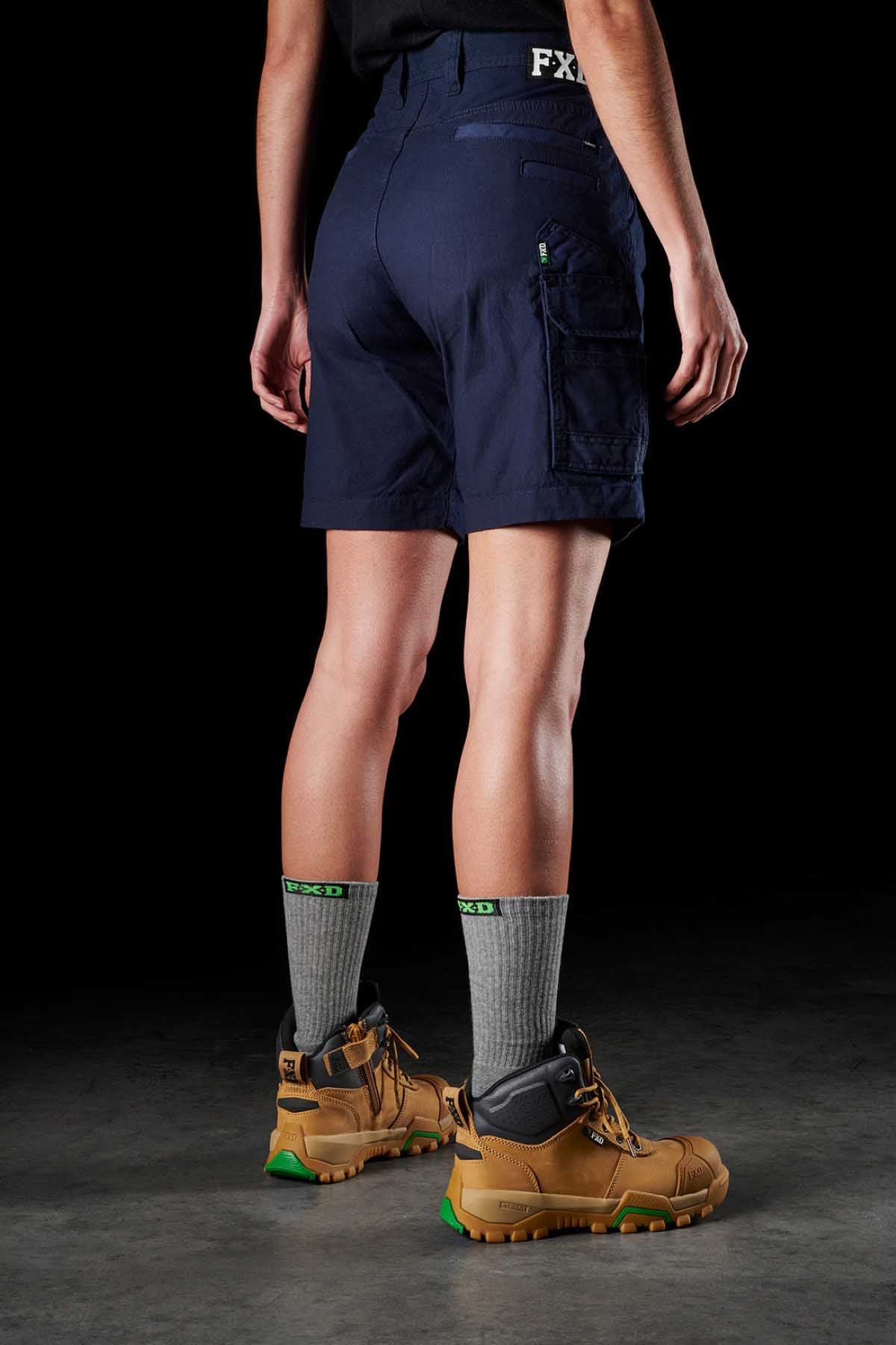The image features a person from the waist down standing on a dark gray floor, donning navy blue FXD Womens WS-3W Shorts equipped with DURA500 reinforced pockets. They are also wearing gray socks featuring a green logo and tan hiking boots accented with green details. Their hands rest naturally at their sides against a black backdrop.