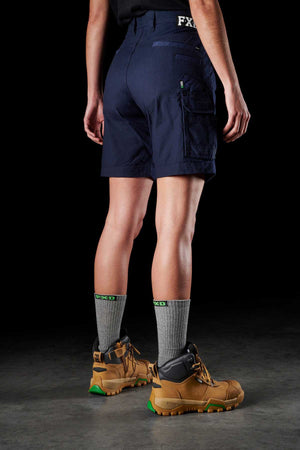 The image features a person from the waist down standing on a dark gray floor, donning navy blue FXD Womens WS-3W Shorts equipped with DURA500 reinforced pockets. They are also wearing gray socks featuring a green logo and tan hiking boots accented with green details. Their hands rest naturally at their sides against a black backdrop.