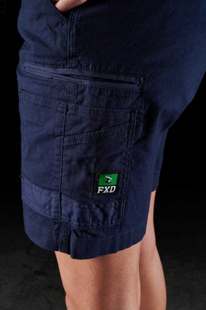 A close-up image of a person's upper thigh and hip area wearing dark blue FXD Womens WS-3W Shorts, which are stretch work shorts equipped with multiple pockets. The utility cargo shorts feature a small green and white FXD logo patch on one of the DURA500 reinforced side pockets. The background is dark.