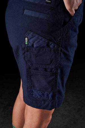 Close-up of a person wearing FXD Womens WS-3W Shorts in navy blue, featuring triple needle seams and multiple pockets, including a prominent one with a green FPPO tag. The background is dark, and only the lower torso and upper legs of the person are visible.