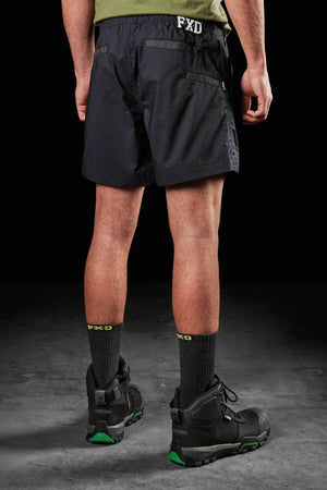 A person stands facing away, wearing a green shirt and FXD WS-4 Shortโblack shorts with the letters "FXD" on the waistband, featuring triple needle seams and made from recycled polyester stretch ripstop fabric. Black socks with "FXD" written in green, along with black work boots featuring green detailing, complete the look. The background is dark.