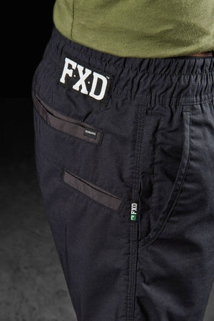 Close-up of a person wearing black FXD WS-4 Shorts made from Recycled Polyester Stretch Ripstop, featuring a comfort stretch elastic waistband with the FXD logo prominently displayed. The shorts have triple needle seams, and one pocket is secured with a zipper while the other with a button, paired with a green shirt tucked in.