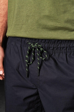 Close-up of a person wearing FXD WS-4 Shorts with a comfort stretch elastic waistband and a green T-shirt. Only the torso and part of the arm are visible. The drawstring on the shorts is green with black and white accents, tied in a bow, showcasing meticulous triple needle seams.