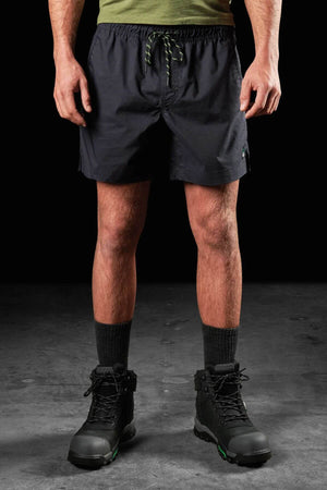 A person standing against a dark background, wearing a green shirt, FXD WS-4 Short with a drawstring and Comfort stretch elastic waistband, tall black socks, and black hiking boots. Only the lower half of their body is visible.