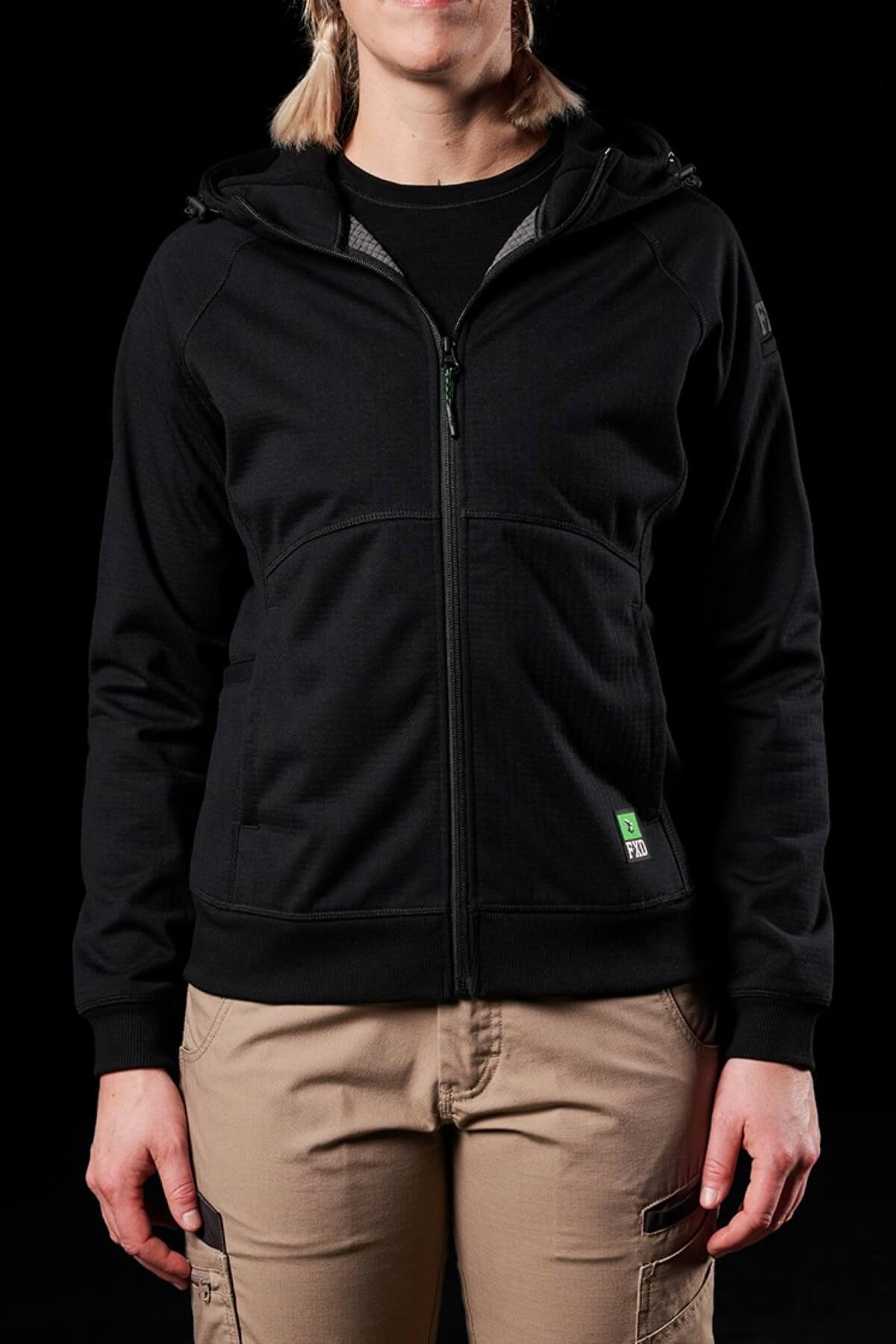 Against a black backdrop, the person wears a sleek FXD Womens WF-3W Jacket. This black zip-up hoodie features a hood, paired with tan cargo pants. Their relaxed stance highlights the jacket's windproof comfort and water-resistant versatility, exuding confidence and style.