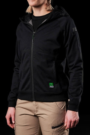 A person in a black FXD Womens WF-3W Jacket and beige cargo pants stands against a dark background. The windproof hoodie, featuring "FXD" on the chest and sleeve, has a hood, with hands resting by their sides.