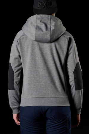 A person stands with their back to the camera, wearing a grey FXD Women's WF-3W Jacket that features black patches on the elbows and a black cap with "FXD" written on it. The dark background makes their outfit stand out. They are also wearing dark blue pants that complement their FXD Women's WF-3W Jacket.