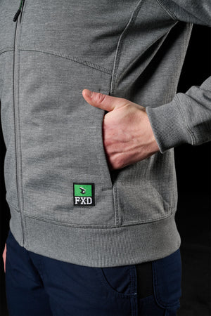 A person is dressed in an FXD Women's WF-3W Jacket, gray with a small green and black "FXD" logo near the pocket. Their right hand is casually placed in the jacket’s pocket, and they are wearing dark blue pants. The black background highlights the clothing's details, including its subtle windproof features.