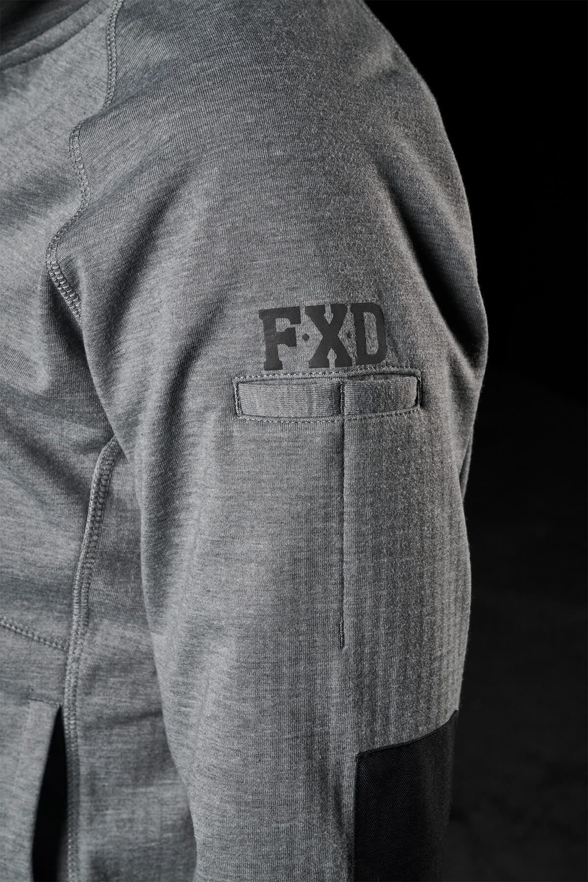 Close-up of a person’s gray long-sleeve shirt sleeve, showcasing the black embroidered FXD logo on the upper arm. The sleeve also features black elbow patches and a small pocket, reminiscent of the design elements found in the FXD Womens WF-3W Jacket. The background is dark.