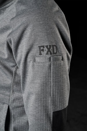 Close-up of a person’s gray long-sleeve shirt sleeve, showcasing the black embroidered FXD logo on the upper arm. The sleeve also features black elbow patches and a small pocket, reminiscent of the design elements found in the FXD Womens WF-3W Jacket. The background is dark.