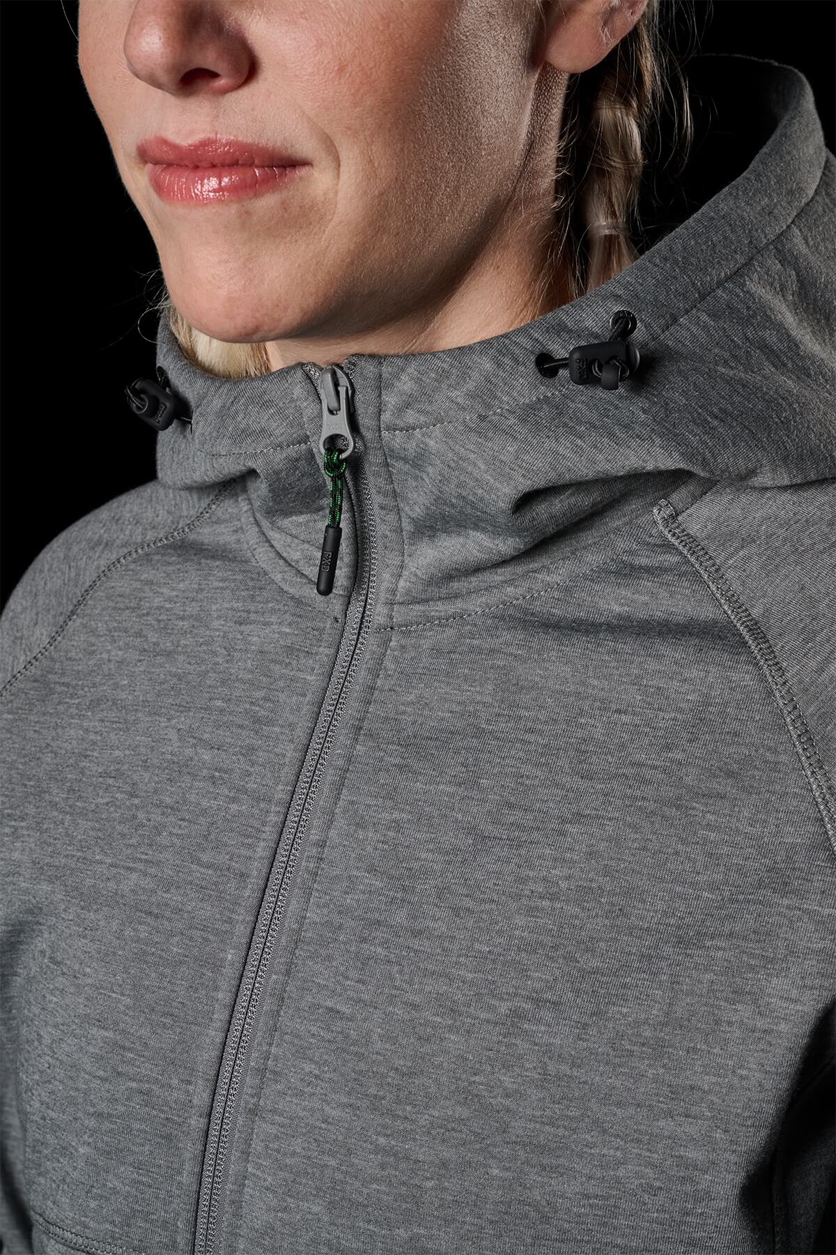 Close-up of a person wearing the FXD Womens WF-3W Jacket in light gray, zipped up to the neck. The windproof jacket features adjustable drawstrings with black toggles on the hood and provides water resistance for added protection. Only the lower half of the person's face is visible against a black background.