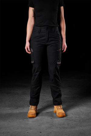 A person stands against a dark background, wearing black FXD Womens WP-7W cargo pants, a black shirt, and tan work boots. The person's arms are by their sides, and their face is not visible.