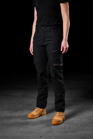 A person wearing a black T-shirt, FXD Womens WP-7W pants in black flex double weave, and tan work boots stands against a dark background. The person has one visible tattoo on their left forearm. Only the lower torso and legs are visible.