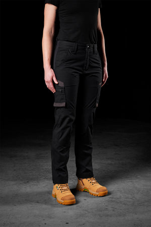 A person wearing a black t-shirt and FXD Womens WP-7W pants with flex double weave and 50+ UPF protection, standing on a dark, textured background. The individual is also sporting tan work boots with yellow laces, presenting a rugged and practical look. Their face is not visible.