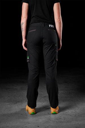 A person wearing black FXD Women's WP-7W cargo pants and tan shoes is shown from the back. The pants, crafted with flex double weave fabric and offering 50+ UPF protection, feature side and back pockets adorned with an "FXD" label. Dressed in a black shirt, they have a tattoo on their right forearm. The background is dark.