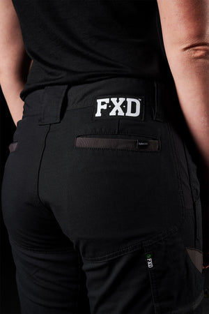 A person is wearing black FXD Womens WP-7W pants, embroidered with the "FXD" logo on the waist, shot from behind. The pants showcase pockets and detailed stitching and are crafted from flexible double weave fabric. They also don a black top, making their attire stand out against the dark background.