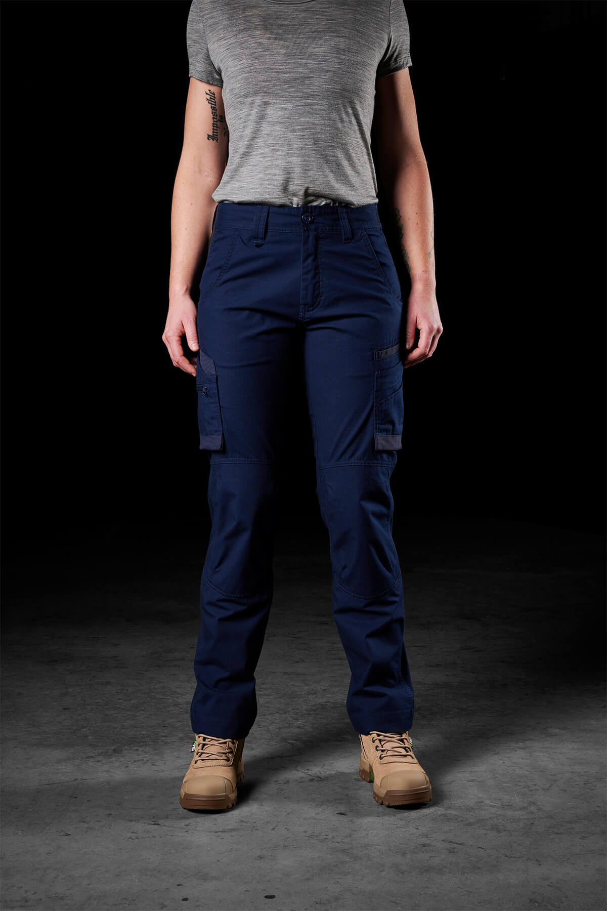 A person wearing a gray short-sleeve shirt, FXD Womens WP-7W pants with flex double-weave hip panels, and tan boots stands against a dark background. The image focuses on the clothing and footwear, as the individual's head is not visible.