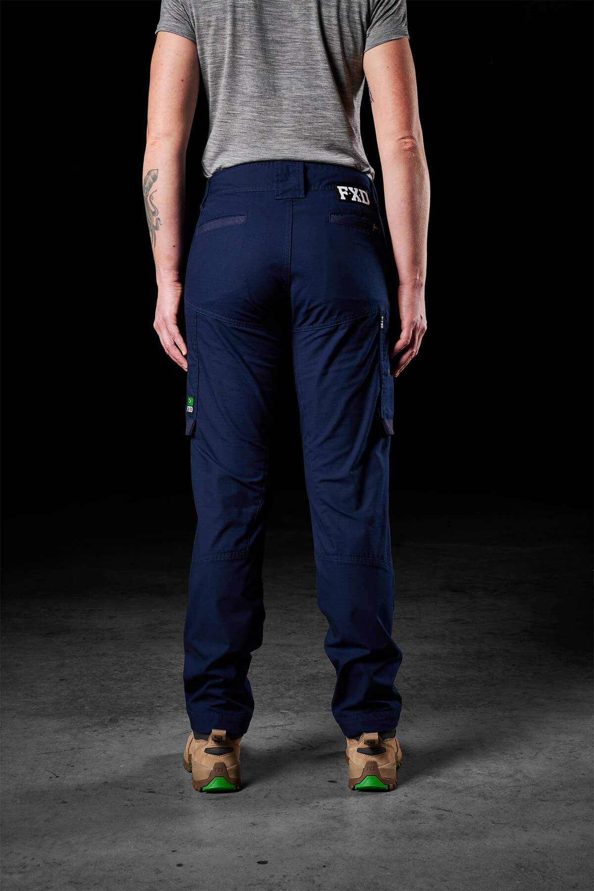 A person stands with their back to the camera, clad in a gray short-sleeve shirt and dark blue FXD Womens WP-7W pants featuring multiple pockets and flex double weave hip panels. They have on beige work boots with green accents. The background is dimly lit, and the ground seems to be concrete.