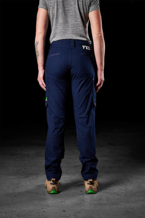 A person stands with their back to the camera, clad in a gray short-sleeve shirt and dark blue FXD Womens WP-7W pants featuring multiple pockets and flex double weave hip panels. They have on beige work boots with green accents. The background is dimly lit, and the ground seems to be concrete.