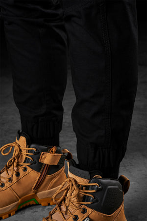 Close-up of a person wearing FXD Womens WP-8W black pants tucked into tan, high-top hiking boots with orange laces. The rugged boots feature zip and lace closures, while the lightweight stretch ripstop pant has elastic cuffs for mobility and comfort. The background is a dark, textured surface.