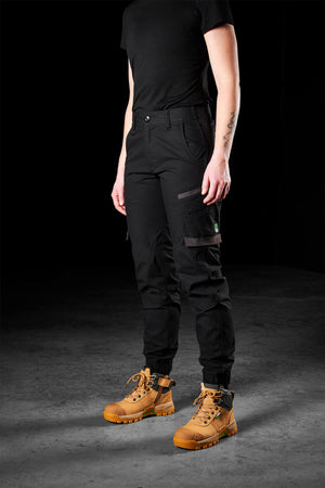 A woman dressed in the FXD Womens WP-8W shirt and lightweight stretch ripstop pants stands on a dark, textured floor. The outfit is paired with tan work boots that have dark laces and rugged soles. Her right arm is visible, showing a tattoo. The background is black.