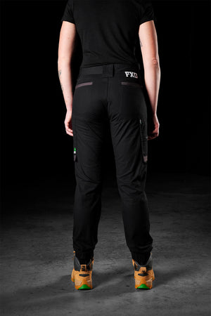 A person wearing black FXD Womens WP-8W workwear pants with abrasion panels and a black T-shirt is standing with their back to the camera. The pants have multiple pockets, and the person has light skin and short hair. They are also wearing light brown, green-soled work boots. The background is black.