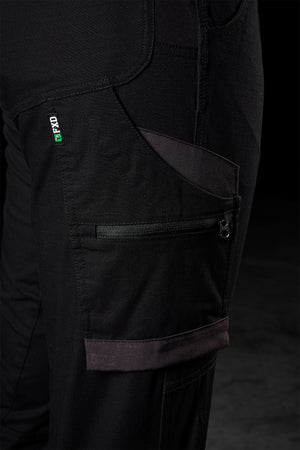 Close-up of a black zippered utility pocket on the FXD Womens WP-8W lightweight stretch ripstop pants, showcasing the detailed stitching and fabric texture. The pants feature additional small pockets and a small green and white tag with text near the pocket seam. The background is dark and out of focus.