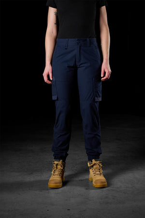 A person stands facing the camera, wearing a black top, FXD Womens WP-8W pants in blue, and tan-colored tactical boots. The dark background highlights the clothing's 50+ UPF protection and the sturdy footwear.