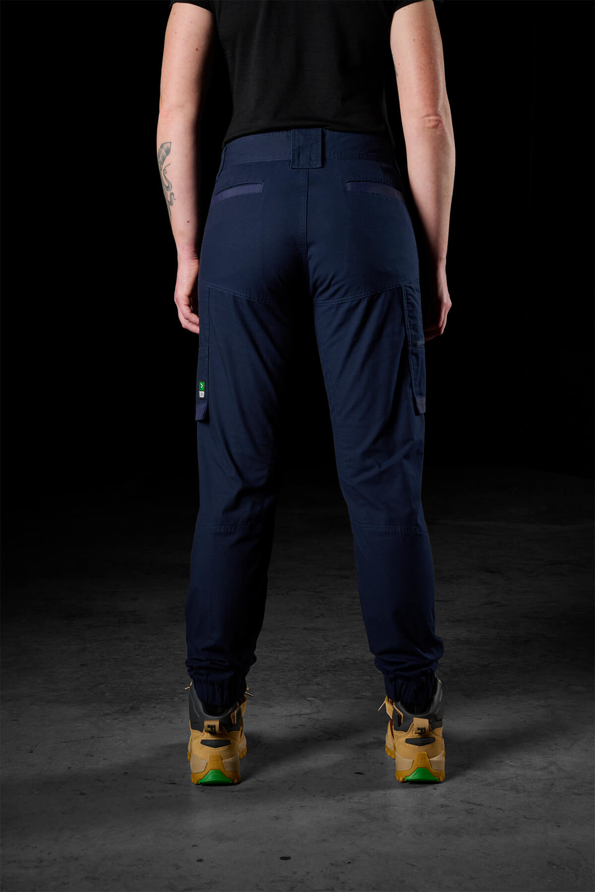 A person stands facing away, highlighting FXD Womens WP-8W stretch ripstop pants with side pockets. They are dressed in a black shirt and tan work boots, with a tattoo visible on their left forearm. The background is dimly lit.