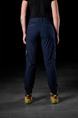 A person stands facing away, highlighting FXD Womens WP-8W stretch ripstop pants with side pockets. They are dressed in a black shirt and tan work boots, with a tattoo visible on their left forearm. The background is dimly lit.