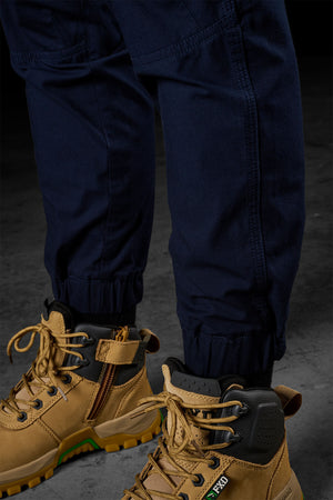 Close-up of a person wearing FXD Womens WP-8W stretch ripstop pants with elastic cuffs, paired with tan work boots featuring black and yellow accents. The boots have visible laces and side zippers. In the background, there is a dark, textured surface.