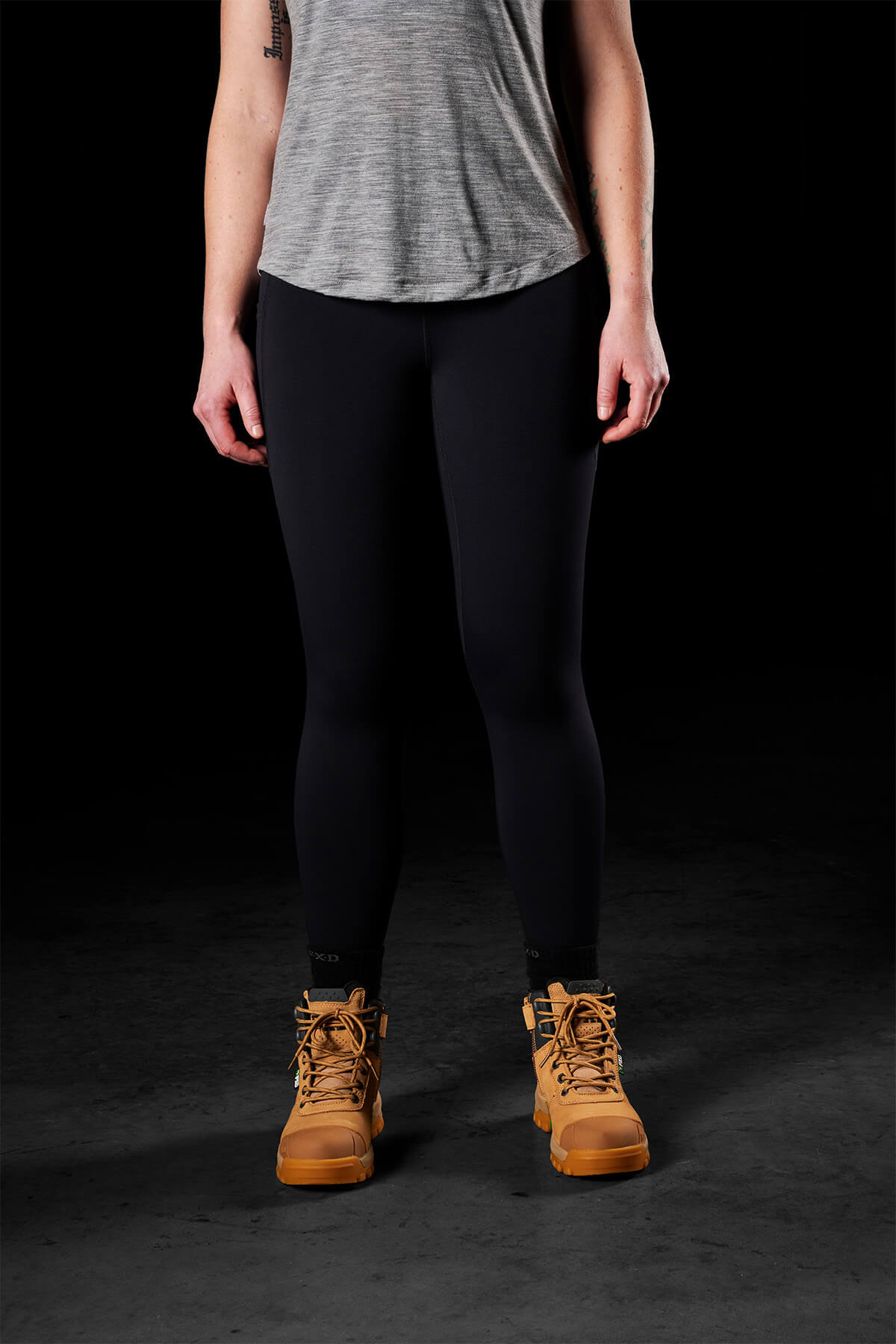 A person wearing a gray shirt, FXD Womens WP-9W leggings with a high-rise waistband, and brown work boots stands against a black background. The image is cropped to show from the shoulders down, concealing the person's face.