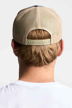 Rear view of a person wearing a beige mesh FlexFit American Stitch Trucker Cap with an adjustable snapback strap and a white shirt. The neutral, plain background accentuates the modern headwear style.