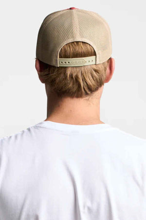 A person wearing a FlexFit American Stitch Trucker Cap with a snapback strap, short blonde hair visible underneath, and a white t-shirt stands facing away from the camera against a plain background.