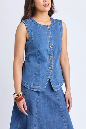 A woman models the Foxwood Abbott Vest in a modern silhouette, paired with a matching denim skirt and chunky bracelets on her left wrist, standing against a plain light background.