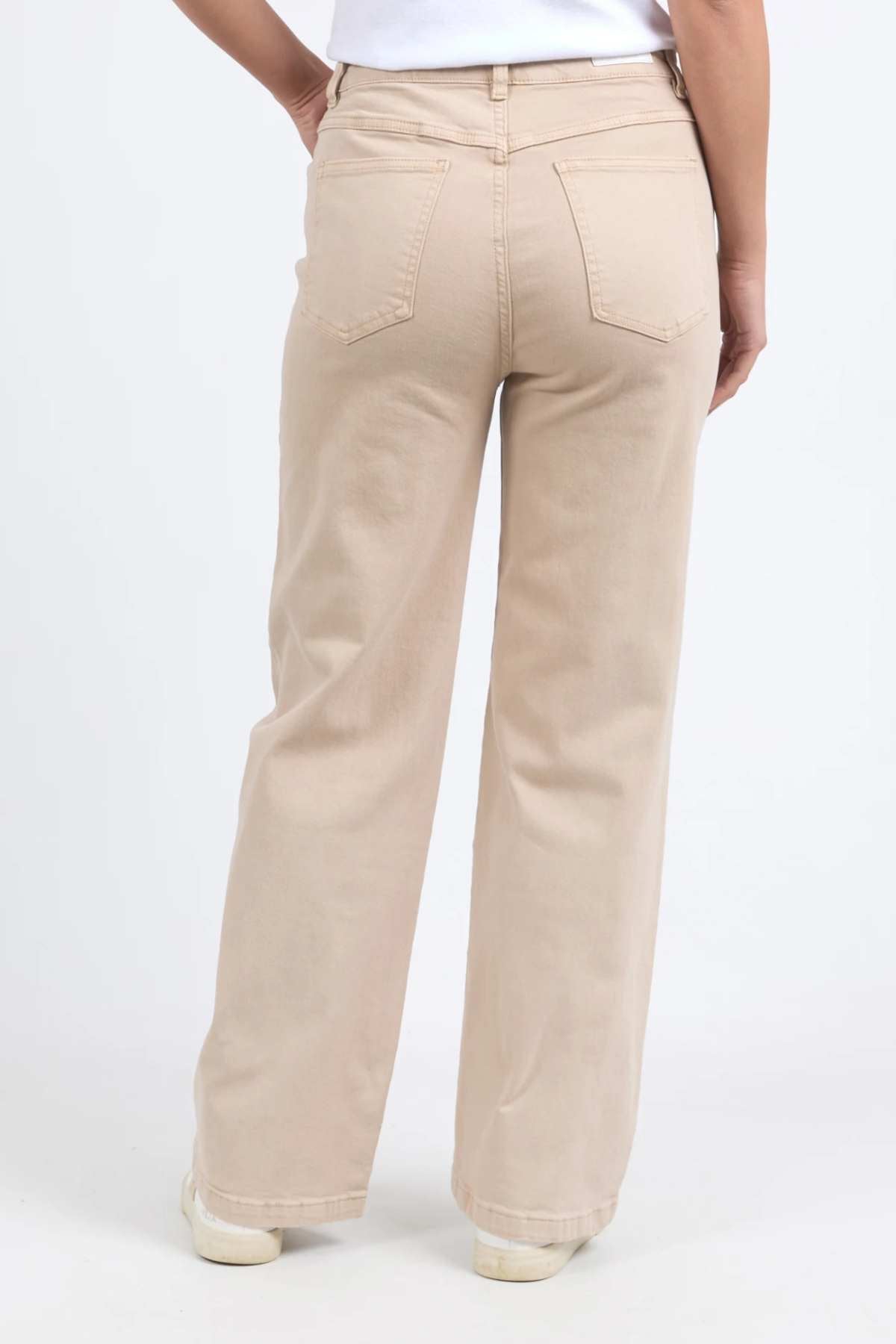 A person wearing the Foxwood Adira Jean in a light wash, high-waisted, stretch denim paired with a white top, viewed from behind. The wide-leg fit of the jeans emphasizes their silhouette as they stand with hands on hips against a plain white background.