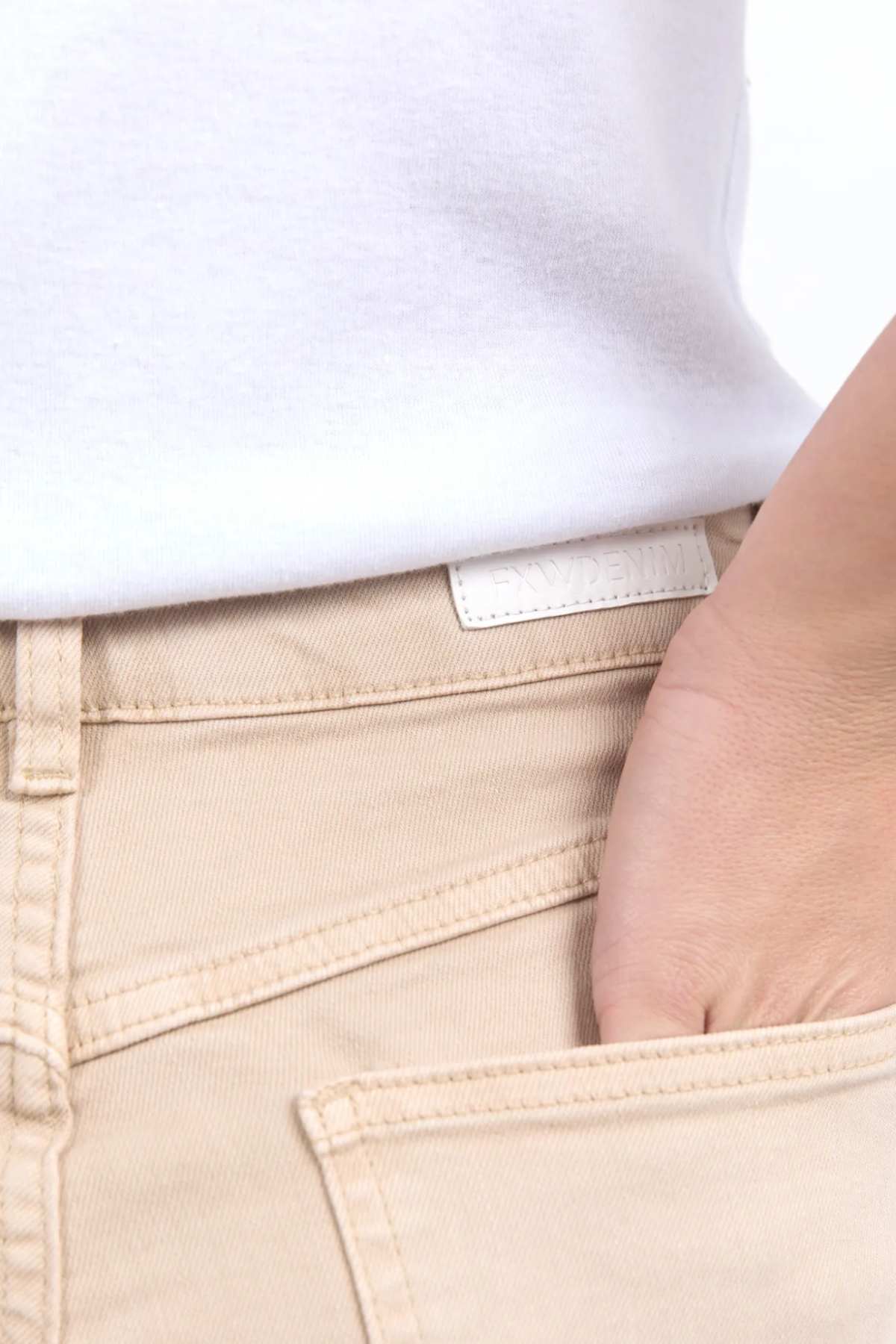 Close-up of a person wearing light beige Foxwood Adira Jeans and a white shirt. Their hand is casually placed in the front pocket of the stretch denim pants, which feature a small white tag on the waistband near the top edge of the pocket.