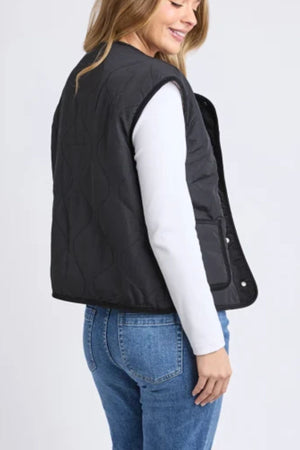 A woman with long, light brown hair is seen from the side and back, wearing the Foxwood Alberta Vest in black with sherpa lining over a white long-sleeve shirt and blue jeans. She is smiling and looking downward.